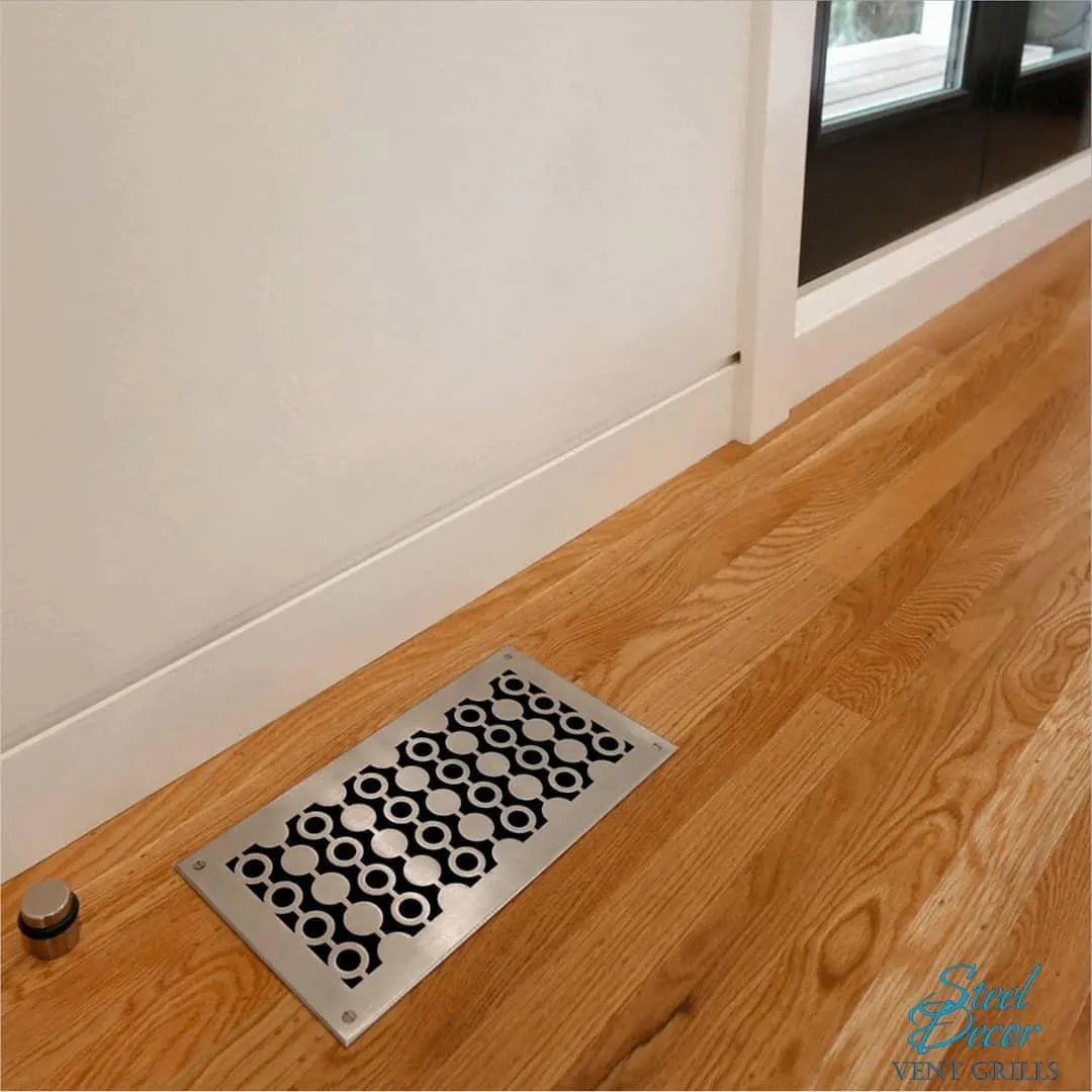 Decorative metal floor vent on a wooden floor with a neutral wall background