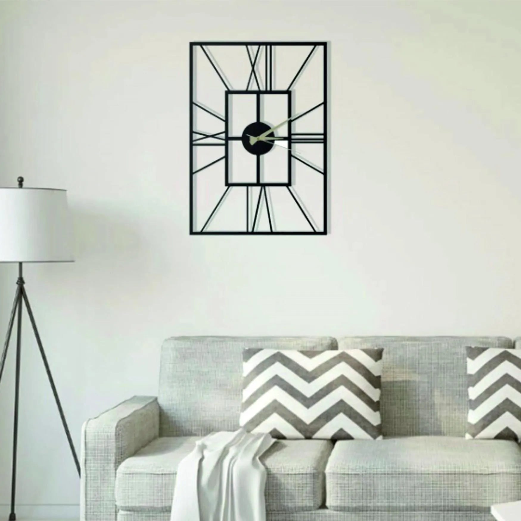 Black geometric wall clock on a white wall above a gray sofa with patterned cushions.
